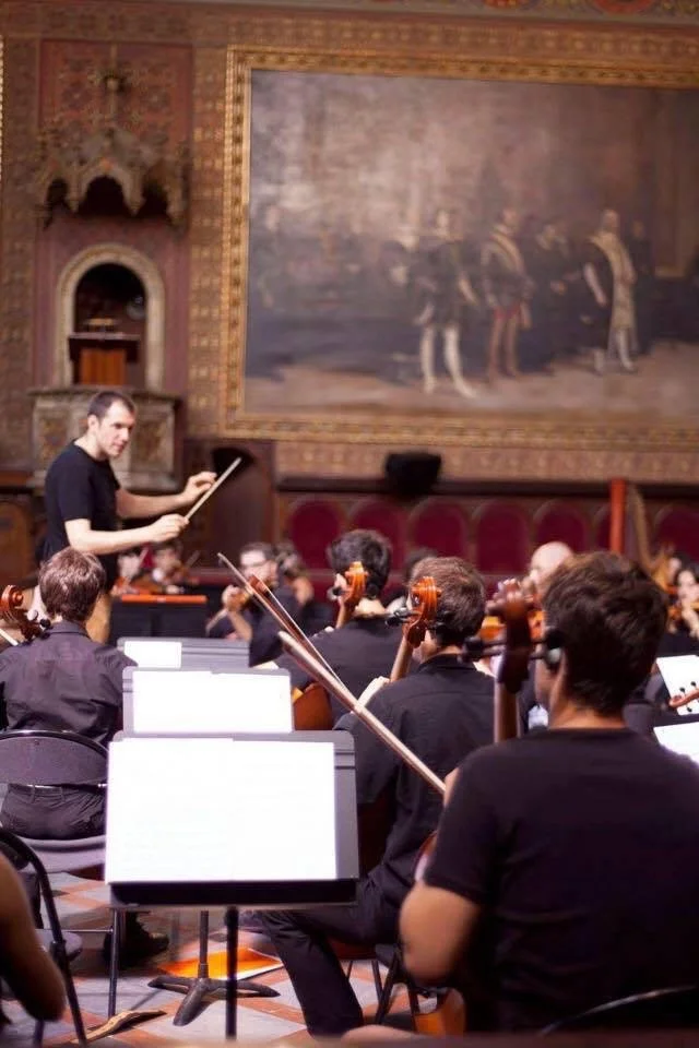 An orchestra rehearsing in a hall with wooden-paneled walls and a large painting in the background, conducted by Gerard Pastor in a black short-sleeve T-shirt. It’s a dress rehearsal.