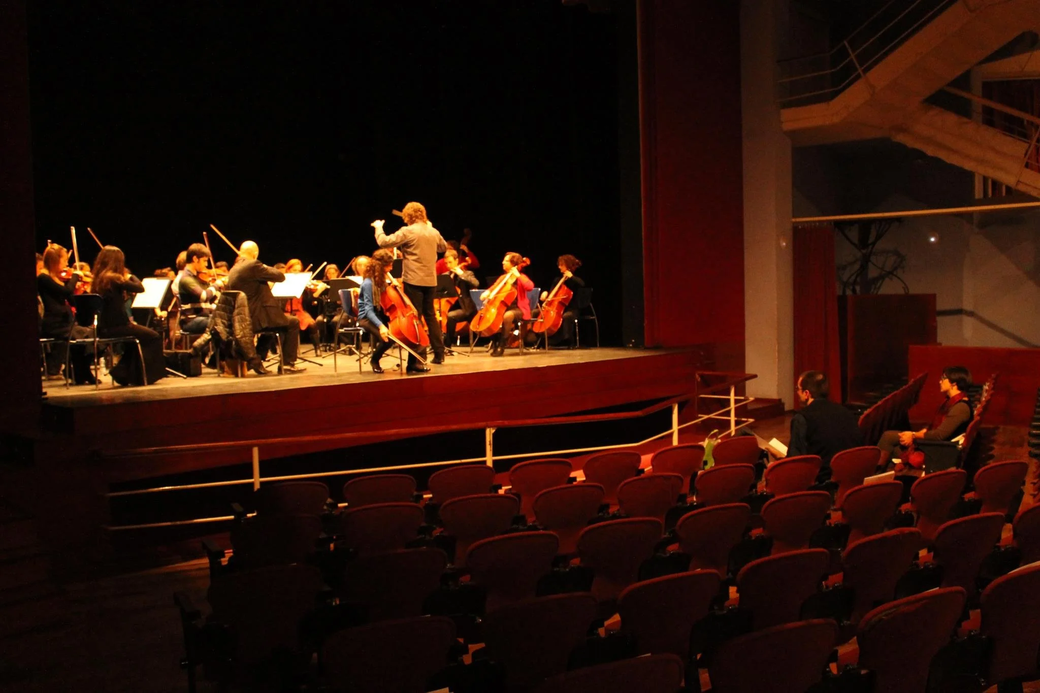 Tomàs Grau conducting a rehearsal of the Franz Schubert Filharmonia with cellist Mariona Camats in a theatre. In the audience, Gerard Pastor and Anna M. Bofarull can be seen.