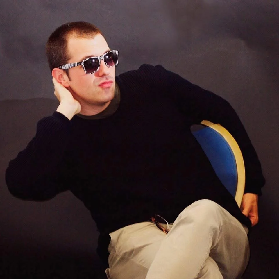 Gerard Pastor sitting humorously on a chair, looking to the side, wearing relatively small sunglasses.