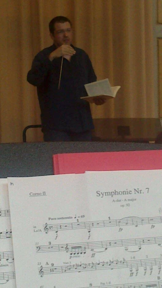 Photograph taken from a musician’s stand during a rehearsal, with Gerard Pastor conducting in the background.